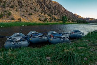 Several inflatable rafts are lined up on the grassy bank of a serene river, with steep rocky hills in the background. The area is lush and green near the water, transitioning to dry, rugged terrain further away. The scene is captured during what appears to be early morning or late afternoon, indicated by the soft, angled lighting.