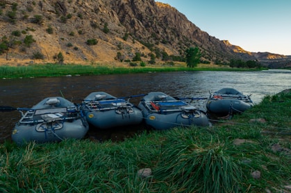 Several inflatable rafts are lined up on the grassy bank of a serene river, with steep rocky hills in the background. The area is lush and green near the water, transitioning to dry, rugged terrain further away. The scene is captured during what appears to be early morning or late afternoon, indicated by the soft, angled lighting.