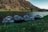 Several inflatable rafts are lined up on the grassy bank of a serene river, with steep rocky hills in the background. The area is lush and green near the water, transitioning to dry, rugged terrain further away. The scene is captured during what appears to be early morning or late afternoon, indicated by the soft, angled lighting.