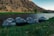 Several inflatable rafts are lined up on the grassy bank of a serene river, with steep rocky hills in the background. The area is lush and green near the water, transitioning to dry, rugged terrain further away. The scene is captured during what appears to be early morning or late afternoon, indicated by the soft, angled lighting.