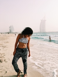 A woman in patterned swimwear stands on a sandy beach, with gentle waves lapping at her feet. Two modern, sail-shaped skyscrapers are visible in the background on a hazy day. Other people can be seen enjoying the water and shoreline.
