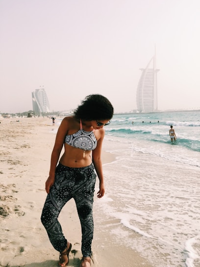 A woman in patterned swimwear stands on a sandy beach, with gentle waves lapping at her feet. Two modern, sail-shaped skyscrapers are visible in the background on a hazy day. Other people can be seen enjoying the water and shoreline.