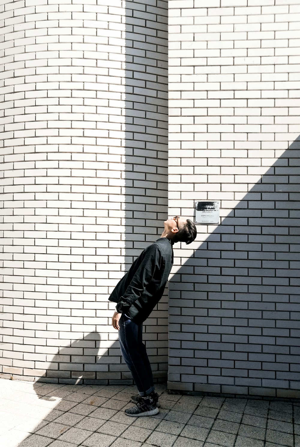 man in black jacket and blue denim jeans leaning on brown brick wall