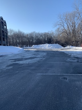 An empty parking lot with a clear, paved surface and patches of snow. A large pile of snow is visible at the far end, surrounded by a line of bare trees without leaves. To the left, a multi-story building with arched balcony openings is present. A clear blue sky stretches above the scene, with a few scattered clouds.