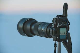 A professional camera with a large telephoto lens is mounted on a tripod. The camera's screen displays an image in focus. The background is a gradient of soft blue and pink, suggesting an open sky during sunrise or sunset.