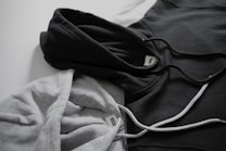 Two hoodies in contrasting colors lie on a flat surface, one gray and the other black. Both have drawstrings visible, and a tag can be seen near the neckline of each hoodie.