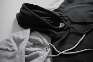 Two hoodies in contrasting colors lie on a flat surface, one gray and the other black. Both have drawstrings visible, and a tag can be seen near the neckline of each hoodie.