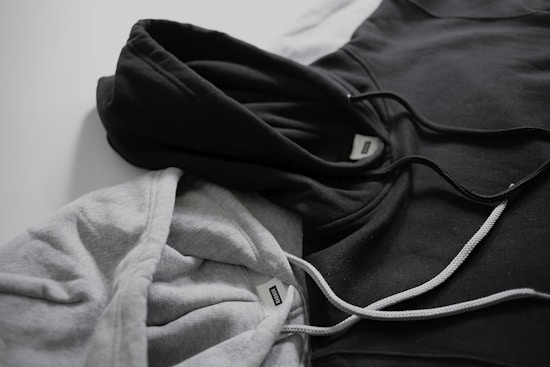 Two hoodies in contrasting colors lie on a flat surface, one gray and the other black. Both have drawstrings visible, and a tag can be seen near the neckline of each hoodie.