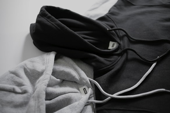 Two hoodies in contrasting colors lie on a flat surface, one gray and the other black. Both have drawstrings visible, and a tag can be seen near the neckline of each hoodie.