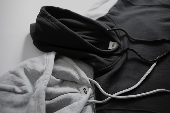 Two hoodies in contrasting colors lie on a flat surface, one gray and the other black. Both have drawstrings visible, and a tag can be seen near the neckline of each hoodie.