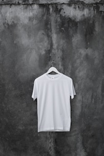A plain white T-shirt hangs on a wooden hanger against a textured, dark, weathered concrete wall.