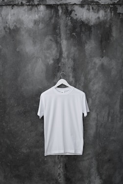 A plain white T-shirt hangs on a wooden hanger against a textured, dark, weathered concrete wall.