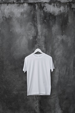 A plain white T-shirt hangs on a wooden hanger against a textured, dark, weathered concrete wall.