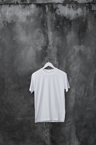 A plain white T-shirt hangs on a wooden hanger against a textured, dark, weathered concrete wall.
