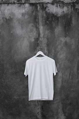 A plain white T-shirt hangs on a wooden hanger against a textured, dark, weathered concrete wall.