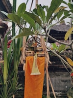 A traditional Balinese offering is placed outdoors, surrounded by tropical plants. The offering basket contains various items like flowers and fruits, set on a stand with orange and yellow fabric draped beneath. The background features greenery and a wooden structure with a thatched roof.