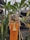A traditional Balinese offering is placed outdoors, surrounded by tropical plants. The offering basket contains various items like flowers and fruits, set on a stand with orange and yellow fabric draped beneath. The background features greenery and a wooden structure with a thatched roof.