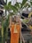 A traditional Balinese offering is placed outdoors, surrounded by tropical plants. The offering basket contains various items like flowers and fruits, set on a stand with orange and yellow fabric draped beneath. The background features greenery and a wooden structure with a thatched roof.
