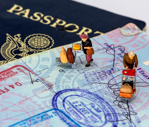 A close-up of a passport with various visa stamps and signatures. On top of the passport pages, there are miniature figures of people depicted as travelers with luggage carts. The background is partially a dark blue passport cover with a golden emblem.