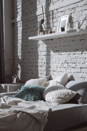 A cozy bedroom featuring a bed with an assortment of cushions and pillows, including a teal fur pillow. Behind the bed is a wall with white exposed brick, casting light and shadow patterns. A floating shelf holds decorative items such as candles, a framed picture, a small potted cactus, and an ornament. To the left, there is a bedside table with a glass vase containing twigs or dried flowers.