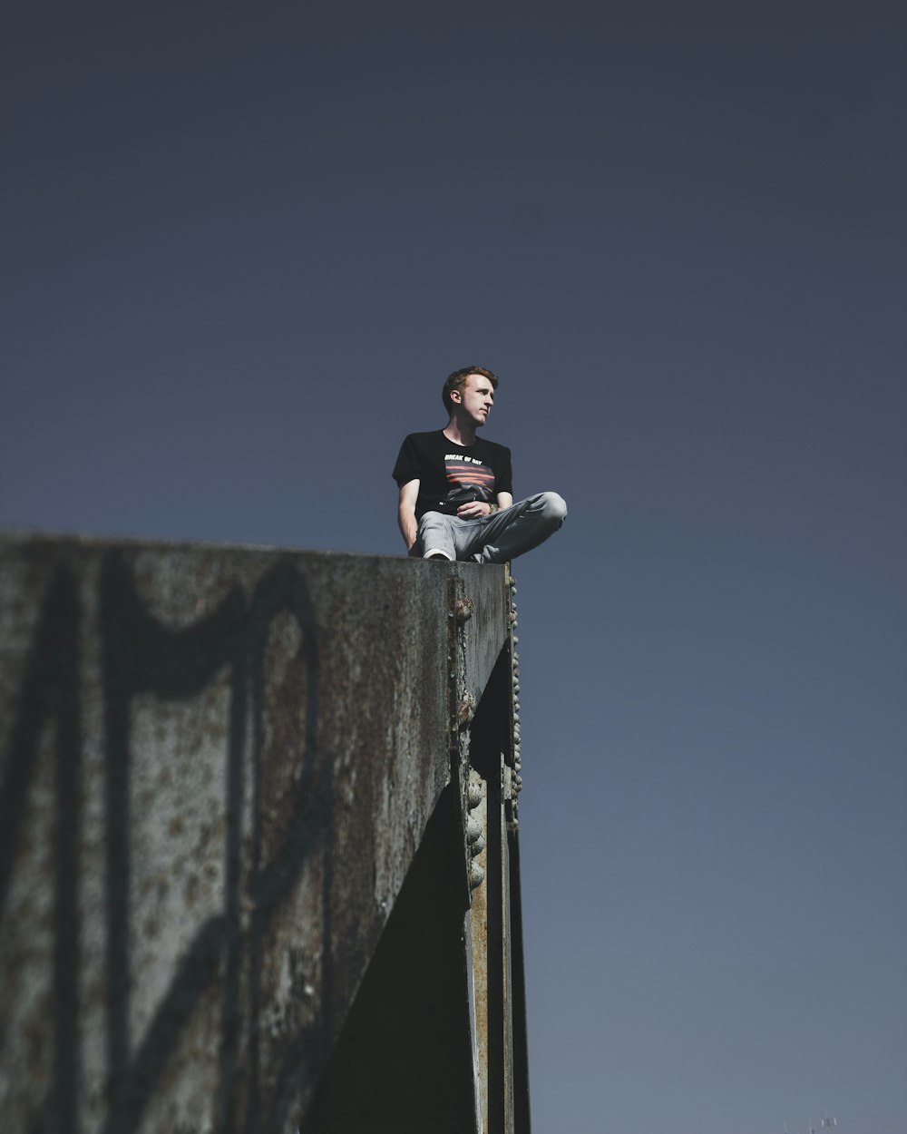 man in black t-shirt and black pants sitting on black metal fence under blue sky