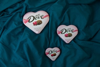 Three heart-shaped Dove chocolate boxes are arranged on a dark teal fabric. The boxes vary in size, featuring a pink and white design with images of chocolate pieces on the lid.