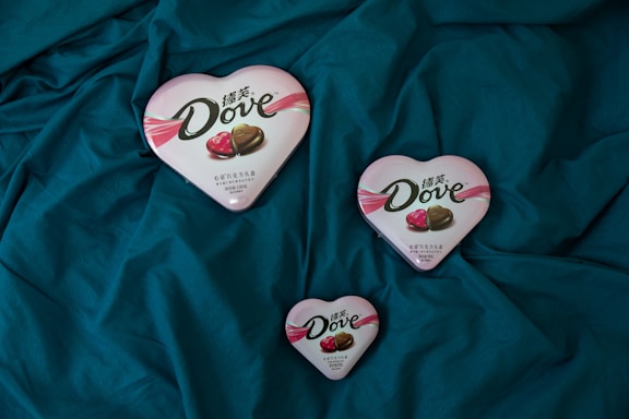 Three heart-shaped Dove chocolate boxes are arranged on a dark teal fabric. The boxes vary in size, featuring a pink and white design with images of chocolate pieces on the lid.