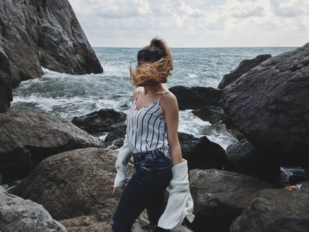 donna in canotta bianca e jeans blu denim in piedi sulla costa rocciosa durante il giorno