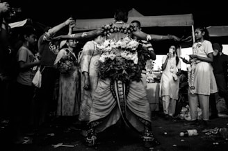 A person dressed in elaborate, traditional attire stands with arms outstretched, surrounded by a group of individuals. The scene appears to be a festive or ritualistic gathering, with people taking photos and some wearing garlands. The image is in black and white, giving it a dramatic and timeless feel.