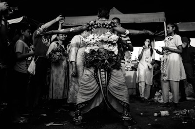 A person dressed in elaborate, traditional attire stands with arms outstretched, surrounded by a group of individuals. The scene appears to be a festive or ritualistic gathering, with people taking photos and some wearing garlands. The image is in black and white, giving it a dramatic and timeless feel.