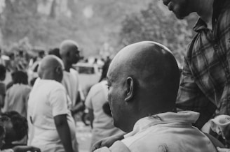 A black and white image of a crowd of people, some with shaved heads, engaged in an outdoor communal activity. The focus is on a person with a freshly shaved head, sitting, possibly receiving a service or participating in a ritual. The setting is outdoors with blurred foliage in the background.