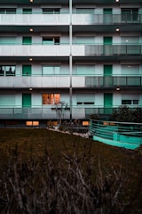 A multi-story residential building with repetitive patterns on the facades, featuring balconies with perforated metal railings. The building is mostly gray and white with teal-colored doors and elements. One window on the first floor is brightly lit from inside. A lawn with a hedge in the foreground provides contrast to the structured architecture.