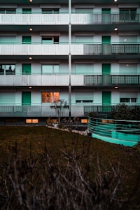 A multi-story residential building with repetitive patterns on the facades, featuring balconies with perforated metal railings. The building is mostly gray and white with teal-colored doors and elements. One window on the first floor is brightly lit from inside. A lawn with a hedge in the foreground provides contrast to the structured architecture.