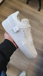 A white sneaker being held in a person's hand, features a sleek design with detailed stitching, a textured sole, and black text on the side. The background shows a wooden floor and the leg of a nearby chair.