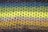 A close-up view of a knitted fabric showcasing rows of openwork stitch patterns. The yarns display a gradient of colors from blues at the top to greens and yellows in the middle, transitioning to browns and grays at the bottom. The texture of the knit is prominent, with the intricate interlocking loops forming a delicate yet vibrant surface.