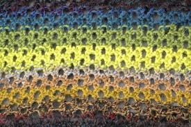 A close-up view of a knitted fabric showcasing rows of openwork stitch patterns. The yarns display a gradient of colors from blues at the top to greens and yellows in the middle, transitioning to browns and grays at the bottom. The texture of the knit is prominent, with the intricate interlocking loops forming a delicate yet vibrant surface.