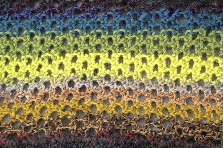A close-up view of a knitted fabric showcasing rows of openwork stitch patterns. The yarns display a gradient of colors from blues at the top to greens and yellows in the middle, transitioning to browns and grays at the bottom. The texture of the knit is prominent, with the intricate interlocking loops forming a delicate yet vibrant surface.