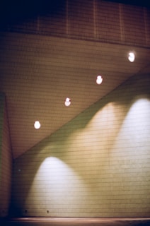 A minimalist interior space featuring a slanted ceiling with four evenly spaced recessed lights. The walls are tiled with a grid-like pattern, and the lighting creates a soft glow on the wall's surface.