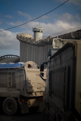 A tall concrete barrier with a watchtower, partially surrounded by a mesh fencing. In the foreground, there are two large trucks, one with a blue tarp covering its load, situated near the wall. The scene takes place under a partially cloudy sky.