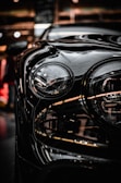 A close-up of a car headlight with smooth, glossy reflections of light on the vehicle's surface. The background appears to be an indoor setting with ambient lighting, giving off a modern and sleek appearance.