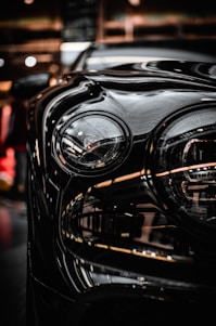 A close-up of a car headlight with smooth, glossy reflections of light on the vehicle's surface. The background appears to be an indoor setting with ambient lighting, giving off a modern and sleek appearance.