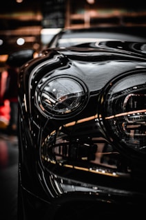 A close-up of a car headlight with smooth, glossy reflections of light on the vehicle's surface. The background appears to be an indoor setting with ambient lighting, giving off a modern and sleek appearance.