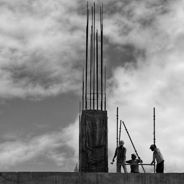 Workers equipped with helmets are engaged in construction work, handling scaffolding and iron rods. The scene is black and white, capturing the imposing structure against a backdrop of cloud-filled sky.