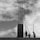 Workers equipped with helmets are engaged in construction work, handling scaffolding and iron rods. The scene is black and white, capturing the imposing structure against a backdrop of cloud-filled sky.