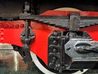 A close-up view of a mechanical component, possibly from a train or heavy machinery. It features a glossy red painted surface, black metal parts with visible bolts, and a textured finish. The detailed view showcases the intricate connections and engineering design.