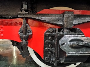 A close-up view of a mechanical component, possibly from a train or heavy machinery. It features a glossy red painted surface, black metal parts with visible bolts, and a textured finish. The detailed view showcases the intricate connections and engineering design.