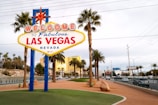 A brightly colored welcome sign for Las Vegas stands prominently in the foreground, surrounded by tall palm trees with a backdrop of a clear blue sky and distant buildings. The iconic sign features bold red and blue lettering with a large star on top, capturing the vibrant essence of the city.