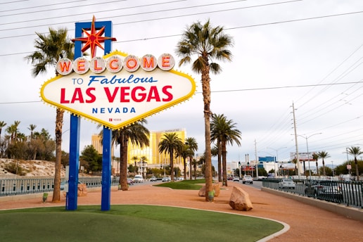 A brightly colored welcome sign for Las Vegas stands prominently in the foreground, surrounded by tall palm trees with a backdrop of a clear blue sky and distant buildings. The iconic sign features bold red and blue lettering with a large star on top, capturing the vibrant essence of the city.