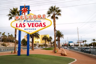 A brightly colored welcome sign for Las Vegas stands prominently in the foreground, surrounded by tall palm trees with a backdrop of a clear blue sky and distant buildings. The iconic sign features bold red and blue lettering with a large star on top, capturing the vibrant essence of the city.