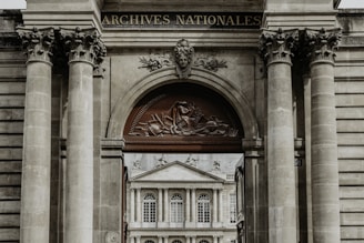 A grand architectural structure with ornate columns and a decorative archway, featuring a relief sculpture and the words 'Archives Nationales'. Through the archway, there is a view of a classical-style building with large windows and ornate detailing.